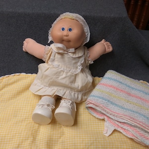 May include: A Cabbage Patch Kid doll wearing a white dress with a lace trim and a white bonnet. The doll is sitting on a yellow and white checkered blanket with a striped blanket beside it.