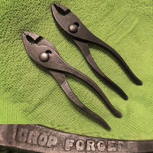 May include: Two dark grey metal slip-joint pliers with serrated jaws and a pivot point. The handles are embossed with the text DROP FORGED. The pliers feature a textured finish and are positioned diagonally on a bright lime green fabric surface.