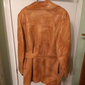 Op de afbeelding: Een bruinleren jas met een patchwork-ontwerp, hangend aan een deur. De jas heeft een riem in de taille en lange mouwen. Het leer heeft een licht versleten uiterlijk.