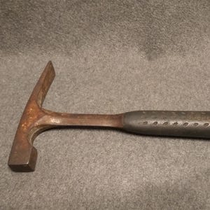 May include: A rusty, vintage rock hammer with a pointed end and a flat, square striking face. The handle is dark gray with a textured grip. This tool is likely used for geological work.