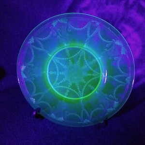 May include: A clear glass plate with a decorative floral pattern, illuminated with a neon green and blue glow. The plate's design includes a central circular motif and a repeating chain-like border.