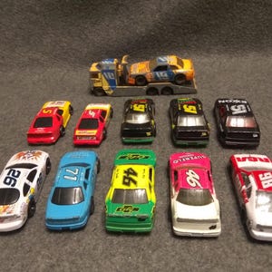 May include: A collection of toy race cars in various colors, including red, yellow, black, green, blue, and white. Some cars have racing numbers and sponsor logos. A car hauler truck is also present, carrying a race car.