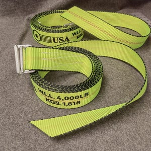 May include: A yellow and black nylon strap with a silver buckle. The strap has the text "WLL 4,000LB KGS 1,818" printed on it. The strap is made in the USA.