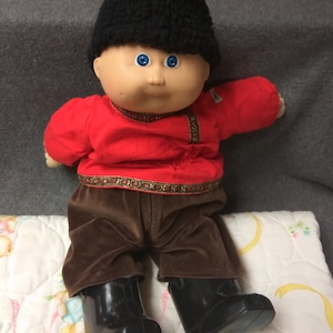 Op de afbeelding: Een Cabbage Patch Kid pop met een zwarte bontmuts, een rood shirt met gouden versiering, een bruine broek en zwarte laarzen. De pop heeft blauwe ogen en een roze teint.