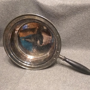 Peut inclure: Une poêle ronde de couleur argentée avec une longue poignée noire. La poêle a un intérieur réfléchissant et la poignée est fixée avec une pièce métallique. La poêle est probablement utilisée pour la cuisine.