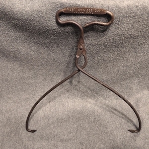 Puede incluir: Una herramienta de pinzas para hielo de metal marrón vintage con un mango en forma de T con el texto en relieve "WEST END ICE CO" y "DULUTH MINN" en la parte superior. La herramienta tiene dos brazos curvos y puntiagudos con bisagra.