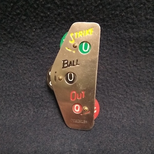 May include: A stainless steel baseball strike zone indicator with the words "Strike", "Ball", and "Out". The indicator shows "0" for each category with green, black, and red colored dials. The word "STAINLESS" is printed at the bottom.