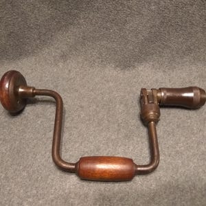 Puede incluir: Un taladro manual vintage con mango y pomo de madera. El taladro es de metal y madera, con un acabado marrón oscuro. La herramienta está diseñada para taladrar manualmente y tiene una apariencia clásica y antigua.