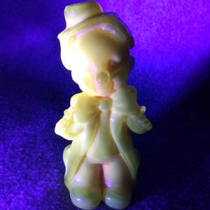 Puede incluir: Una figura amarilla vintage de una persona con sombrero y abrigo. La figura tiene una pajarita y está de pie. La figura está sobre un fondo púrpura.