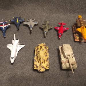 May include: A collection of toy military vehicles and aircraft, including fighter jets, a space shuttle, and tanks. The toys are painted in various colors, such as blue, red, yellow, and camouflage patterns.