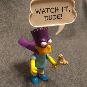 May include: A Bart Simpson figurine with a purple cape, hat, and a slingshot. The figure wears a blue shirt, shorts, and shoes. A speech bubble above the figure reads "WATCH IT, DUDE!"