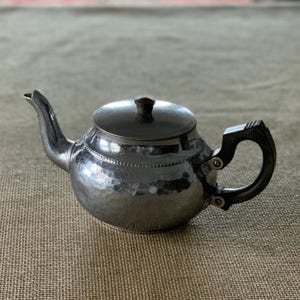 Puede incluir: Una pequeña tetera plateada con textura martillada. Tiene un pico curvo, un asa articulada y una tapa con un pequeño pomo. El asa es oscura y la tetera está sobre una superficie de color neutro. Es probable que sea de metal.