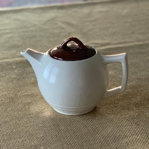 May include: A white ceramic teapot with a brown lid and a handle. The teapot is sitting on a brown textured surface.