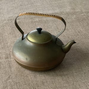 May include: A vintage brass teapot with a rounded body, a curved handle with a woven rattan grip, and a small spout. The lid is brass with a black knob. The teapot is a warm, antique brass colour.