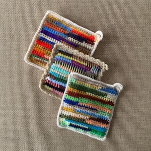 May include: Three handmade crochet pot holders in various sizes. Each pot holder features a colorful striped pattern with a white or beige border. The pot holders have a loop for hanging. The color palette includes red, blue, green, yellow, and purple.