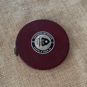 May include: A maroon-colored Favorite Wyteface 100 ft measuring tape by Keuffel & Esser Co. The tape measure has a circular design with a silver metal center and a metal clasp on the side.