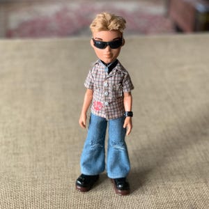 Könnte beinhalten: Puppe mit blonden Haaren und Sonnenbrille, trägt ein kariertes Hemd, blaue Jeans und schwarze Schuhe. Die Puppe hat eine schwarze Uhr am Handgelenk. Das Hemd hat einen roten Aufnäher.