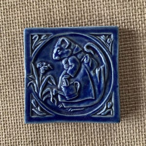 May include: Square, cobalt blue ceramic tile with a relief design. The design features a stylized depiction of a cat-like figure holding a pitcher, surrounded by floral elements within a circular frame. The tile has a glossy finish.