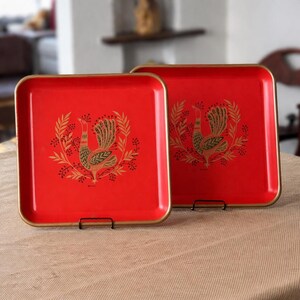 May include: Two square red trays with gold edging. Each tray features a gold and black rooster design surrounded by a wreath. The trays are displayed on a neutral-coloured surface.