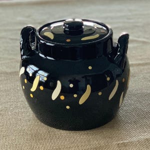 Può includere: Un vaso di ceramica nero con coperchio, due manici e decorazioni dorate e bianche. Il vaso ha una finitura lucida e una forma arrotondata, adatto per la cucina o la decorazione.