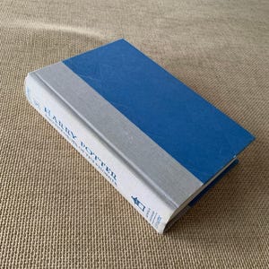 May include: A hardcover book titled "Harry Potter and the Order of the Phoenix." The book has a blue cover and a gray spine with white text. The book is lying on a textured, tan surface.