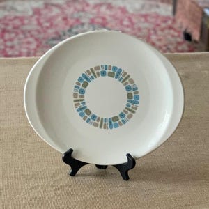 Peut inclure: Une assiette en céramique blanche avec un bord surélevé, ornée d'un motif central de formes géométriques bleues, vertes et beiges disposées en cercle. L'assiette est présentée sur un support noir, mettant en valeur son esthétique vintage.