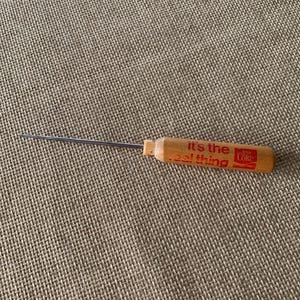 Puede incluir: Un picahielos vintage con mango de madera. El mango es de color marrón claro y presenta el logotipo de Coca-Cola y la frase "it's the real thing" en rojo. La punta metálica es plateada y se extiende desde el mango.