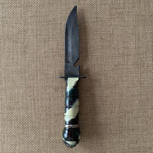 May include: A black survival knife with a serrated blade and a camouflage handle. The knife has a bottle opener and a guard. The handle is green, black, and white.
