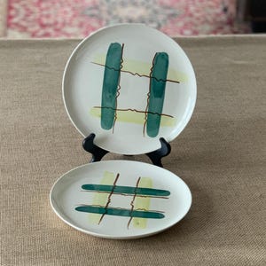 Könnte beinhalten: Zwei weiße Keramikteller mit modernem Design aus der Mitte des Jahrhunderts. Jeder Teller hat ein grünes und gelbes Gittermuster mit braunen Akzentlinien. Die Teller werden auf schwarzen Ständern präsentiert.