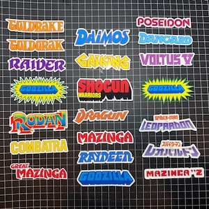 May include: A collection of colorful die-cut stickers featuring various retro anime and superhero titles. Titles include "Goldrake", "Godzilla", "Mazinga", "Spider-Man", and more, in bold, stylized fonts with white borders.