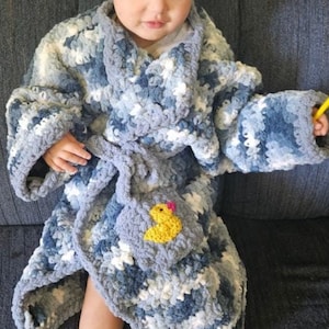 May include: A young child wearing a blue and white crocheted robe with a yellow rubber ducky patch. The robe has a belt and is made of a soft, fluffy yarn.