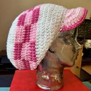 Può includere: Un berretto all'uncinetto con un motivo a scacchi in tonalità rosa chiaro e rosa scuro, contrastato da sezioni in bianco solido. Il cappello presenta una corona arrotondata e una visiera corta e rigida con un bordo rosa scuro in filato.