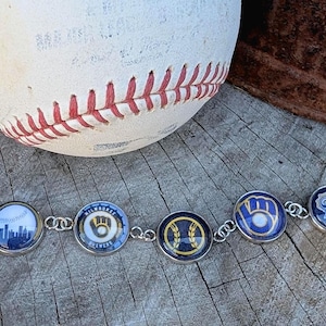 May include: A silver-toned link bracelet featuring seven circular charms with various Milwaukee Brewers baseball team graphics, including the state of Wisconsin, a city skyline, team logos, and a mascot, displayed against a white baseball.