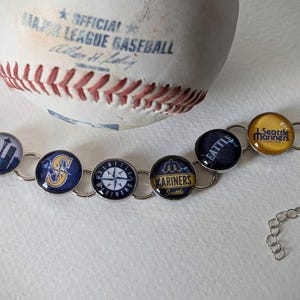 May include: A silver bracelet featuring circular charms with various Seattle baseball team logos and designs. A baseball rests in the background. The bracelet has a chain closure.