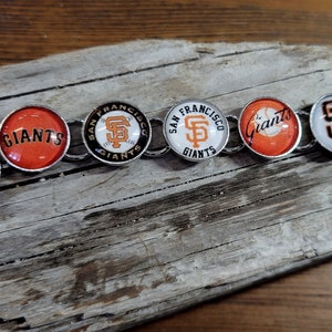 May include: A silver chain bracelet with five round charms featuring the San Francisco Giants logo. The charms are white with orange and black accents. The charms alternate between the team's logo and the word "Giants".