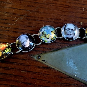 May include: A silver chain bracelet with round glass charms featuring images of classic horror movie characters, including Dracula, Frankenstein, and the Creature from the Black Lagoon.
