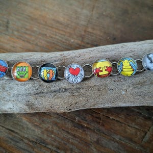 May include: A silver-toned bracelet featuring a series of round, colorful charms with various designs. The charms include a red heart, cartoon figures, and abstract art. The bracelet is displayed on a piece of weathered wood.