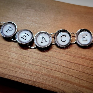 May include: A silver charm bracelet with round, silver-toned charms featuring the word "PEACE" in black lettering, peace signs, and other designs.