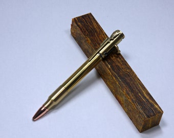 May include: A gold-toned pen made from a bullet casing with a copper tip. The pen is lying on a piece of wood with a brown and orange marbled pattern.