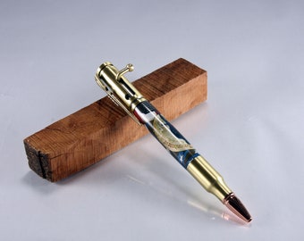 May include: A gold-toned pen made from a bullet casing with a blue, white and red American flag design and the text "We the People" on the barrel. The pen is resting on a piece of wood.