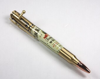 May include: A gold-toned pen with a brass bullet casing design. The pen has a Jack Daniel's Tennessee Whiskey label on the barrel.
