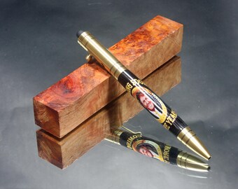 May include: A black and gold pen with a portrait of a man on the barrel. The pen is on a piece of wood with a red and brown grain pattern. The text on the pen reads "Big 80's 7 Years".