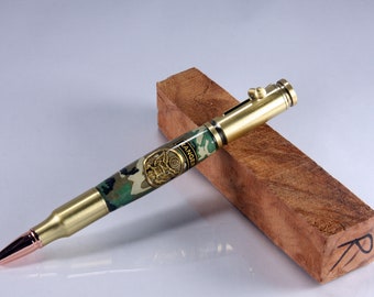 May include: A brass pen with a camouflage pattern and a gold Ranger emblem. The pen is made from a bullet casing and has a copper tip. The pen is resting on a block of wood.