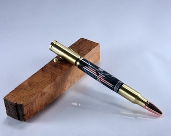May include: A gold and black pen with an American flag design is displayed on a wooden stand. The pen is made from a bullet casing and has a gold-coloured tip.