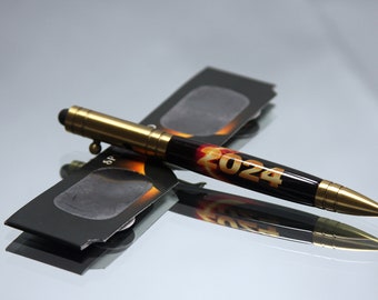 May include: A black and gold pen with the number "2024" printed on it. The pen is lying on top of a pair of black eclipse glasses.