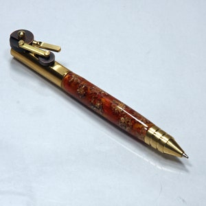 May include: A gold and brown pen with a unique, intricate mechanism at the top. The pen has a wood-like finish with a reddish-brown color and gold accents.