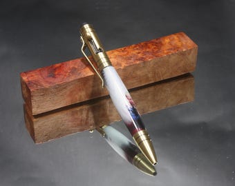May include: A gold and white pen with a wood grain pattern on the barrel. The pen is resting on a piece of wood with a reddish-brown colour.
