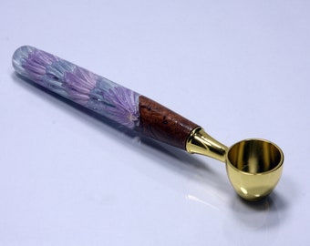 May include: A gold metal coffee scoop with a wooden handle. The handle is decorated with purple and blue flowers embedded in clear resin.