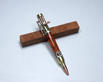 May include: A handcrafted pen with a wooden barrel and metallic accents. The pen has a copper-coloured tip and a brass-coloured top, with intricate details. The pen is displayed on a rectangular wooden block.