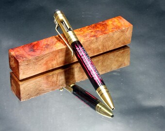 May include: A black and pink pen with gold accents and the words "Air Pistol Off" written on the side. The pen is resting on a piece of wood with a reddish-brown colour.
